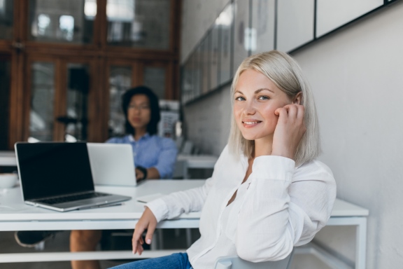 A young, smiling blonde woman dressed in a white blouse and blue jeans sits, relaxed, at a white desk with a laptop open in front of her