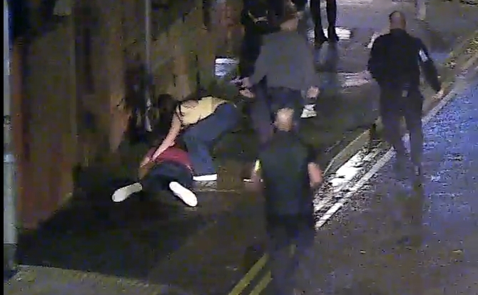 Still picture taken from the CCTV footage showing the victim face down in the street with a woman bending over him in concern and several men running past them in pursuit of Saxton, the perpetrator.